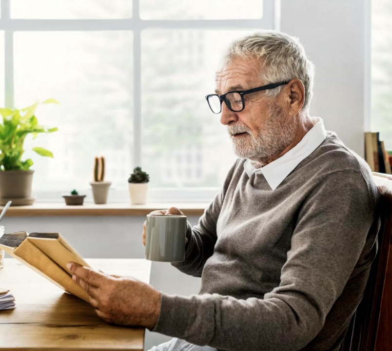 An older person reading at a table with a cup of coffee